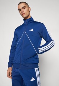Modra zip-up trenirka s visokim ovratnikom, belimi diagonali in tremi belimi črtami na rokavih. Vsebuje Adidas logo.