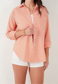 Chemise à rayures avec boutons en corail et blanc, en tissu léger ; présente une coupe décontractée, une poche poitrine et des manches retroussées.