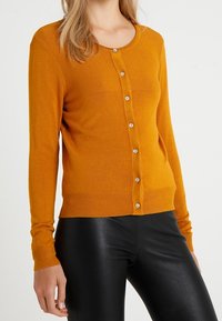 Senapsgul cardigan med rundad halsringning, långa ärmar och knappstängning fram. Har ribbad fåll och mjuk textur.