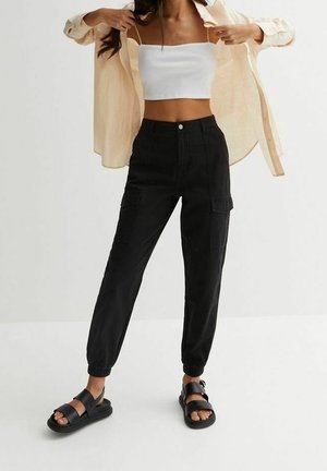 Zwarte cargobroek met elastische boorden en grote zijzakken, gecombineerd met een witte cropped top en zwarte sandalen met bandjes. Lichte beige blouse is opengeknoopt.