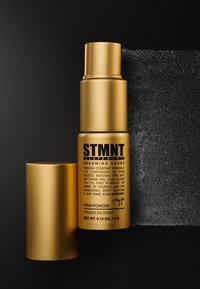 Flacon pulvérisateur en aérosol doré avec un bouchon noir, étiqueté "STMTN Spray Powder" en texte noir. Présente une finition métallique lisse et un fond mat.