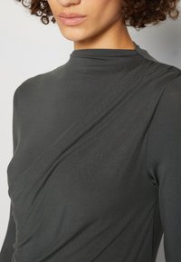 Kvinna i en mörkgrå höghalserad långärmad topp med subtila diagonala sömdetaljer, lockigt hår och naturlig läppglans.