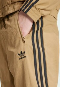 Beige tracksuit pants with elastic waistband, featuring a zippered pocket and black three-stripe design. Adidas logo in black.