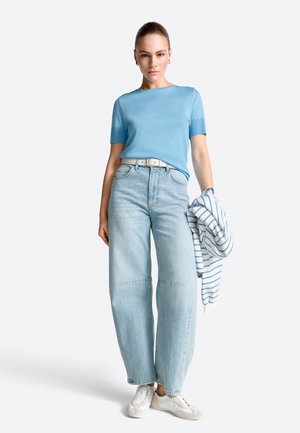 Fiatal nő világoskék rövid ujjú ingben, magasderekú, bőszárú farmernadrágban, fehér sportcipőben, egyik kezében csíkos dzsekit tartva.