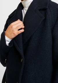 Marineblauwe wollen blendjas met een textuurafwerking, dubbelzijdig ontwerp, grote revers en ronde knopen, gedeeltelijk getoond op een figuur.
