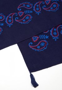Tissu bleu marine avec broderie paisley en rouge et bleu, agrémenté de fleurs. Fini avec un pompon à l'ourlet.