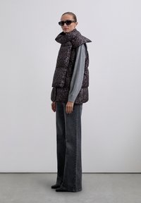 Woman wearing black leopard print puffer vest with attached scarf, gray long-sleeve sweater, dark wide-leg jeans, black shoes, and sunglasses.