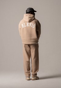 Beige hettegenser med "BLKVIS" logo i hvitt på ryggen, sammen med matchende joggebukser. Materialet ser mykt ut, med en avslappet passform, akkompagnert av joggesko.