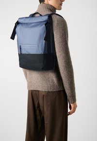 Sac à dos bleu à rabat avec une surface texturée, fond noir, poche frontale zippée, bretelles réglables et design minimaliste.