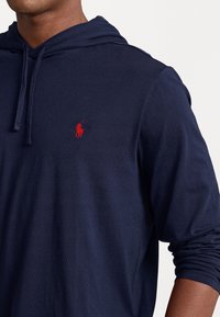 Námořnická modrá mikina s kapucí z lehkého materiálu. Obsahuje kapuci se stahovací šňůrkou a červeným vyšitým logem na levé hrudi. Dlouhé rukávy se manžetami.