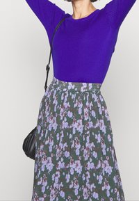 Lila räfflad långärmad topp kombinerad med en grön blommönstrad kjol med ljuslila blommor och en utsvängd design. Axelväska ingår.