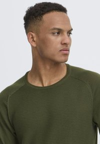 Jersey de color verde oliva, de punto, con cuello redondo y textura de canalé, que presenta mangas raglán y un corte casual. Sin patrones ni acentos visibles.