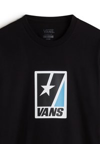 Mustan puuvillapaitojen, joissa on suuri suorakulmainen valkoinen logo tähdellä ja sinisillä yksityiskohdilla. Klassinen istuvuus, jossa on pyöreä pääntie.