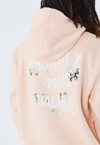 Sudadera de color durazno con capucha, que presenta el texto blanco "PROJECT X PARIS GALERIE" y gráficos florales en la parte posterior. Textura de tela suave.