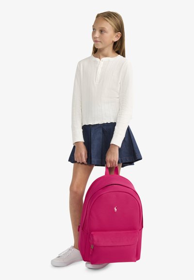 Rucsac roz cu o finisare din material neted, formă rotunjită, buzunar frontal și accent cu logo. Purtați de o persoană îmbrăcată cu un top alb și fustă din denim.