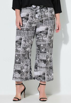 Henkilö, joka käyttää mustavalkoisia kuvioituja lyhytlahkeisia housuja, joissa on solmittava vyötärö, ja mustia korkokenkiä, seisoo valkoista seinää vasten.