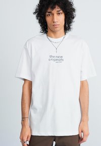 Ung person med lockigt hår iklädd en vit t-shirt med texten "the new originals jazz club", lager på lager av halsband och bruna byxor, stående.