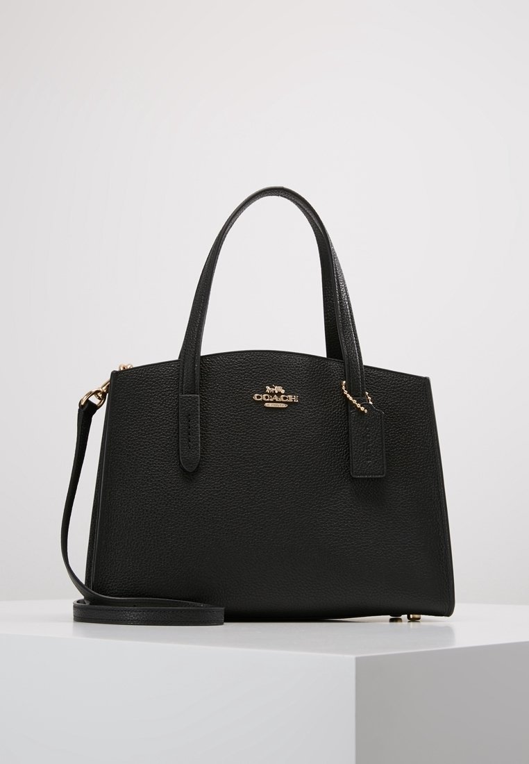Sac à main Coach en cuir noir texturé avec deux poignées, une bandoulière amovible et un logo doré avec une étiquette sur une surface blanche.