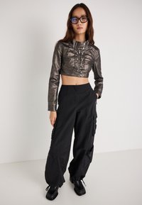 Μεταλλικό κοντό μπουφάν με κουμπιά, συνδυασμένο με μαύρο παντελόνι cargo με τσέπες και ρυθμιζόμενα μανίκια. Χοντρά μαύρα παπούτσια με πλατφόρμα.