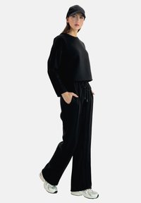 Felpa nera a maniche lunghe e taglio corto, abbinata a pantaloni jogger neri con strisce laterali. Include un cappellino nero e sneakers metallizzate.