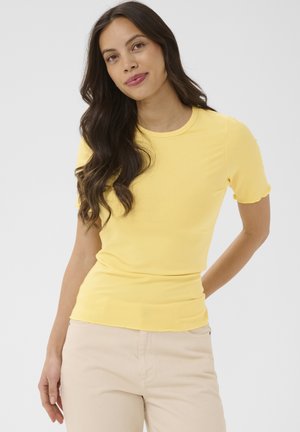 Kobieta o długich ciemnych włosach, ubrana w dopasowany żółty prążkowany top z krótkim rękawem i beżowe spodnie, pozująca na jednolitym jasnym tle.