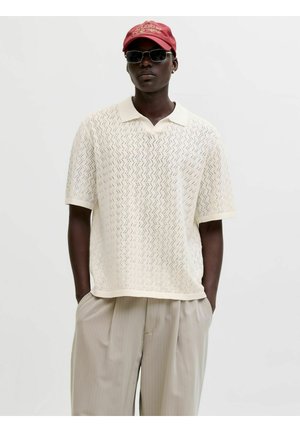 Mężczyzna ubrany w kremowe, dziergane polo z krótkim rękawem, jasno-beżowe spodnie w cienkie paski, czerwoną czapkę z daszkiem i ciemne okulary przeciwsłoneczne, z rękami w kieszeniach.