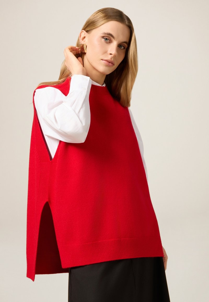 Roter, gestrickter ärmelloser Pullover mit geripptem Saum und seitlichen Schlitzen, übereinander getragen mit einem weißen Hemd mit Knöpfen. Glatte Textur und lockere Passform.