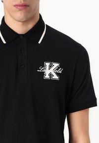Černé polo tričko s bílými akcenty na límci. Na levé hrudi je výrazné vyšívané logo s písmeny "K" a "Lagerfeld".