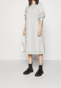 Vestido sudadera gris de gran tamaño con mangas largas, aberturas laterales y puños acanalados, combinado con botas negras de tobillo gruesas y un bolso pequeño blanco.