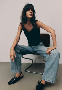 Zwarte mouwloze top, lichtblauwe wijde jeans en glanzende zwarte instappers. Model zit op een stoel met een minimalistische achtergrond.