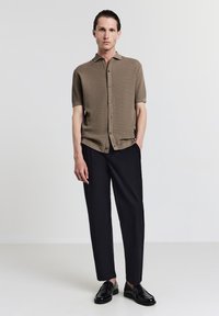 Beige Polohemd mit kurzem Arm, gestrickt mit einem strukturierten Muster, mit Knopfleiste vorne, kombiniert mit lockeren, schwarzen Hosen und glänzenden schwarzen Slippern.