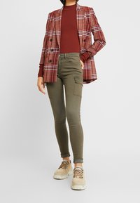 Czerwony w kratę blazer z podwójnym zapięciem na burgundowym topie w prążki. Dopasowane oliwkowe spodnie cargo z kieszeniami i beżowe sneakersy.