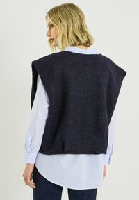 Chaleco de punto azul marino texturizado con amplios orificios para los brazos, usado sobre una camisa a rayas azul claro. Incluye un dobladillo recto y un ajuste relajado.