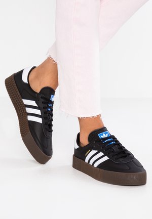 Stopy w czarnych sneakersach Adidas Samba z białymi paskami i brązowymi podeszwami, w połączeniu z jasno-różowymi spodniami z postrzępionymi nogawkami.