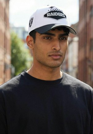 Young man wearing a white Las Vegas Raiders baseball cap and black shirt, standing outdoors in an urban street setting.