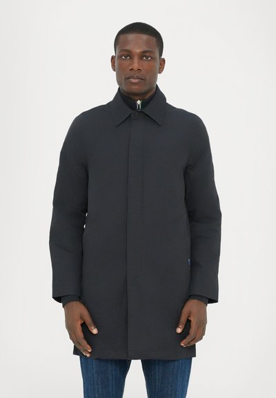 Cappotto nero con taglio dritto, maniche lunghe e chiusura con bottoni. Presenta un colletto a punta e una trama sottile. Abbinato a jeans blu.