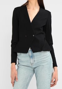 Kvinna klädd i en figurnära svart blazer med två knappar och ljusblå högkortsjeans, stående mot en neutral bakgrund.