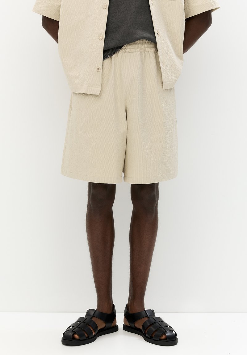 Beige shorts bermuda bertekstur dengan pinggang elastis, dipadukan dengan sandal datar hitam yang memiliki tali silang. Kemeja abu-abu polos dengan lengan pendek.