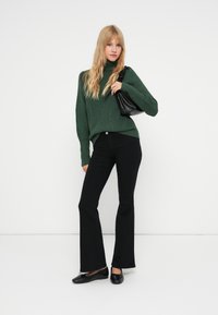 Pullover de gola alta em malha canelada verde, jeans flare pretos e sapatos rasos pretos. Modelo segurando uma pequena bolsa preta, em pé contra um fundo branco.