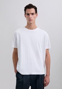 T-shirt blanc à col rond fabriqué en coton lisse ; coupe détendue ; manches courtes ; sans graphiques ni motifs ; associé avec un pantalon sombre.