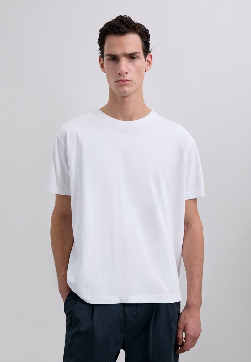 T-shirt blanc à col rond fabriqué en coton lisse ; coupe détendue ; manches courtes ; sans graphiques ni motifs ; associé avec un pantalon sombre.