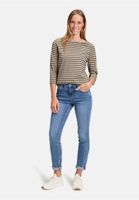 Gestreiftes Langarmhemd in neutralen Tönen, kombiniert mit hellblauen, eng anliegenden Jeans und weißen Sneakers. Lächelndes Model steht mit Händen in den Hüften.