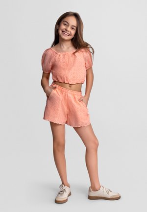 Fille souriante debout les mains dans les poches, portant un crop top en broderie anglaise pêche et un short assorti avec des baskets blanches sur fond gris.