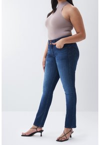 Mujer con vaqueros de tiro alto azules, top sin mangas color malva y sandalias de tacón negras con tiras, de pie con una mano en el bolsillo.