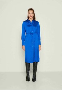 Sieviete stāv vienkāršas fona priekšā, tērpta garām piedurknēm, pogājama zilā kleitā ar jostas auklu un melnām līdz ceļgaliem garām zābakiem.