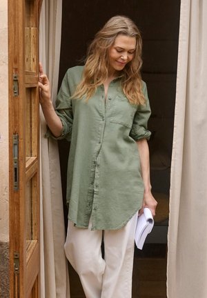 Femme aux longs cheveux ondulés vêtue d'une chemise verte boutonnée et d'un pantalon blanc, debout dans une porte, tenant des papiers roulés, regardant vers le bas et souriant.