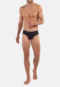 Schwarze Unterhose aus glattem, dehnbarem Stoff mit niedrigem Bund und nahtlosem Gummibund. Das Modell steht barfuß.