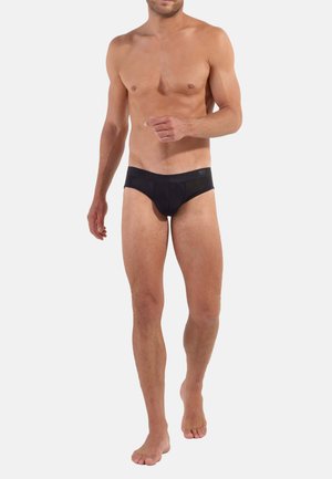 Schwarze Unterhose aus glattem, dehnbarem Stoff mit niedrigem Bund und nahtlosem Gummibund. Das Modell steht barfuß.