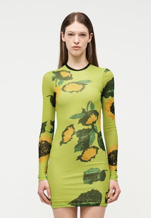 Γυναίκα που φοράει εφαρμοστό lime πράσινο φορέμα με μακριά μανίκια και μεγάλα κίτρινα και πράσινα σχέδια ηλίανθων, στέκεται μπροστά σε ένα απλό φωτεινό φόντο.