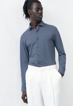 Muž s tmavou pleťou a dredmi nosí modrú vzorovanú košeľu s dlhým rukávom zastrčenú do bielych nohavíc s vysokým pásom, stojaci pri bielej stene.